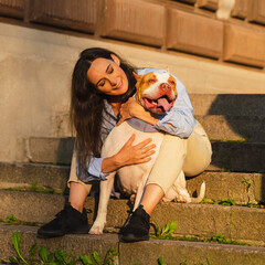 Brunette woman in casual clothes sitting on stone staircase and hugging bicolor white-brown enthusiastic american pitbull terrier dog at dawn. Walking and training happy dog outside