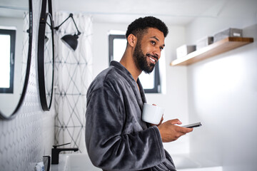 Young man with coffee and bathrobe indoors in bathroom at home, morning routine concept.