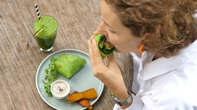 Top View Of Young White Woman Takes A Bite Of Her Delicious Vegan Burrito Served With Sauce, A Side Of Sweet Potato And Refreshing Cold Pressed Green Juice