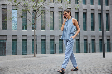 Full length shot of happy positive woman in blue outfit enjoys cellphone conversation in roaming while traveling abroad makes smartphone calling via app strolls outdoor near modern establishment