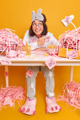 Positive optimistic brunette Asian woman in nightwear works at office desk surrounded by cut paper...