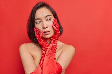 Charming young Asian woman with dark hair keeps hands on face looks tenderly aside stands bare shoulders wears long gloves poses against vivid bright red background with copy space for promotion