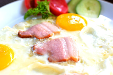 Fried eggs with pieces of bacon, tomatoes and cucumbers. Breakfast on a white plate