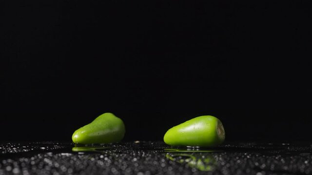 Green hot jalapeno peppers hitting a wet surface bounce upward and form a spray against a black studio background. Wallpaper spicy ripe vegetable for cooking or restaurant. Close up. Slow motion.
