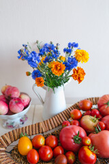 Summer harvest still life, garden flowers in the white jug on the table, wicker dish with ripe tomatoes, vase with apples, rural interior decoration