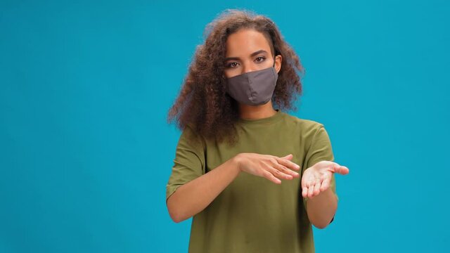 Sign language, deaf African American girl shows hand gesture using sign language. Young woman in protective mask looking at camera wearing in an olive t-shirt. Isolated on blue background.