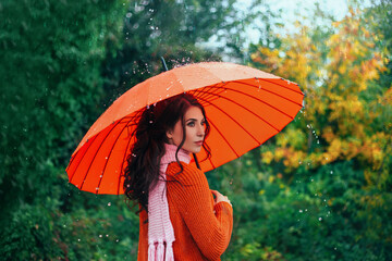 Young beauty woman walks in bad weather under summer rain drops water, holds bright orange red...