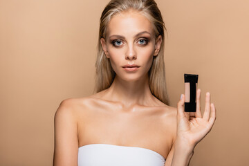 pretty woman with grey eyes and blonde hair showing tube of makeup foundation isolated on beige