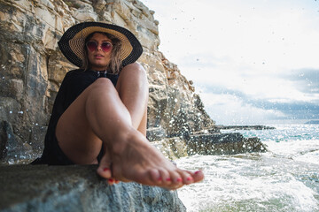 Attractive girl wearing a black dress, sunglasses and a hay hat sitting by the sea. Stormy day by the beach in Croatia, warm summer of 2021. Barefoot feet in foreground
