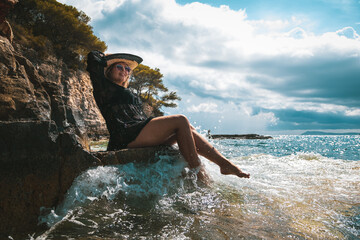 Brunette girl sitting on a rock by the sea on a sunny summer day in Croatia, suprised and shocked by the wave hitting the shore and splashing her. Fun on the beach while traveling in 2021