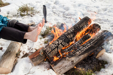 Men bare feet are heated by the campfire, standing next to shoes, in winter snowy day.