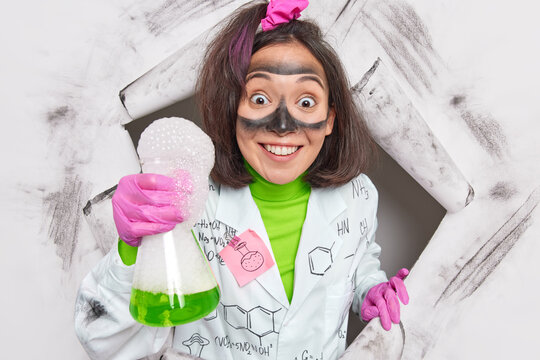 Smiling positive Asian female researcher conducts chemical experiment in laboratory holds glassware with green liquid and bubbles mixes reagents dressed in medical coat breaks through paper wall