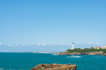 The coastline of the famous Biarritz, France. Ocean waters in the foreground. Lighthouse on the horizon, over a blue sky