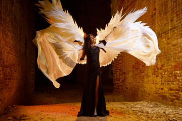 A brunette girl with long hair and white wings in an evening dress in a vintage Gothic castle. Woman who looks like an witch or angel in an abandoned old church. Female Model posing in dark hall