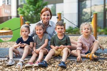 Group of small nursery school children with teacher outdoors on playground, looking at camera.