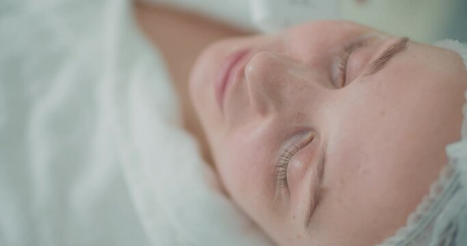 Close-up face of a female client, she is laying with closed eyes while having a cosmetology procedure.