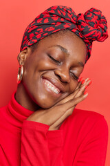 Dark skinned woman keeps eyes closed tilts head hands pressed together near face has gentle expression wears silk scarf tied on head casual turtleneck isolated over red background. Happy emotions