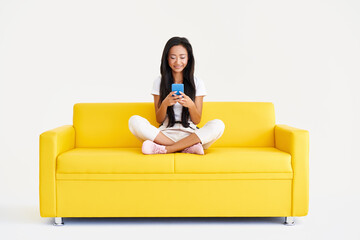 Smiling asian woman types text message, browses internet on cell phone sitting on yellow couch