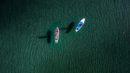 Two sporty peoples are walking on sup boards with paddles at turquoise water. Aerial drone view