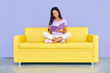 Obraz premium Pretty asian woman reading book sitting at cozy yellow couch, concepts of home and comfort