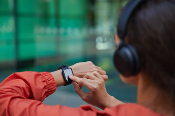 Unknown woman checks results of fitness training on smartwatch listens music via headphones dressed in anorak poses against blurred background. Back view. People health tracking and sport concept