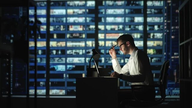 Serious man employee sit at desk develop start up ideas, search problem solution working hard use laptop app at night in modern skyscrapers office room. Deadline, careerism, workaholism concept