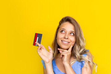 Close-up of a young blonde woman with wavy hair holding a plastic credit card in her hand thinking about how to spend the money looking up at empty copy space isolated on a color yellow background