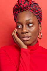 Vertical shot of thoughtful dark skinned young woman keeps hand on face focused aside wears casual turtleneck kerchief tied on head has pensive expression poses against vivid red background.