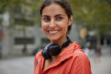 Outdoor shot of happy young dark haired Caucasian woman wears anorak headphones around neck smiles positively poses against blurred background at street walks and has sport training in open air