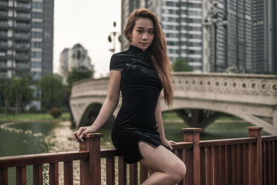 Beautiful young asian woman in black mini dress standing near handrail in the park. Long hair. Nice posture. Looking at camera with copy space. 