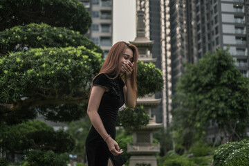 Portrait of beautiful young asian woman in casual black dress standing in the park and laughing. Covers her mouth with her hand. Long hair