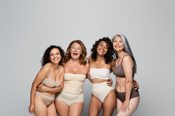 Multiethnic body positive women in lingerie hugging and looking at camera isolated on grey
