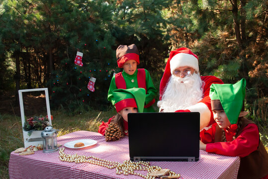 Santa Claus In A Red Suit Sits At A Table With Elves Children Looking Into A Laptop In Summer.
Holiday Advertising Concept For Christmas Or New Year