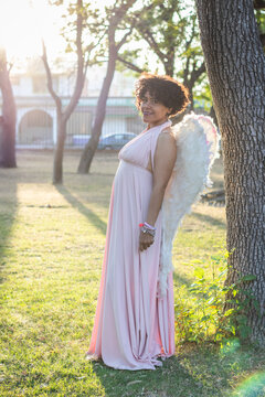 Mid adult afro mexican woman standing in a park, smiling and looking at the camera, dressed in a pink dress and wearing wings like an angel