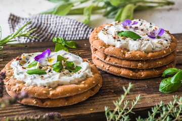 Round crispbread with herbal cream cheese, fresh herbs and edible blossoms on a wooden board