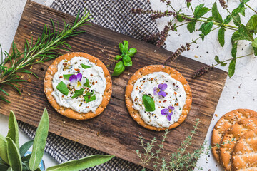 Round crispbread with herbal cream cheese, fresh herbs and edible blossoms on a wooden board, top view