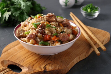 Fried rice with beef, vegetables, garlic, ginger and green onions, sprinkled with sesame seeds on a wooden board on a dark background. Asian cuisine.