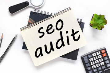 Seo Audit, an open notebook lies on a closed notebook near a calculator and a magnifying glass on a white workspace.