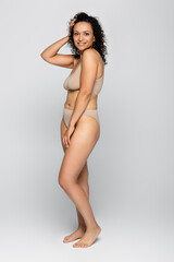 Smiling african american woman in underwear adjusting hair on grey background, body positive concept
