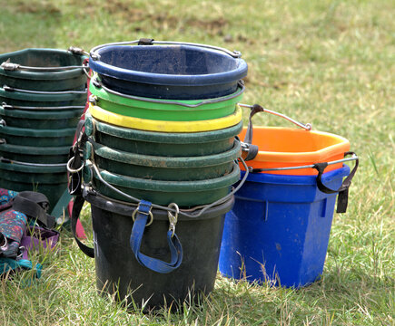 A variety of feed and water buckets stacked in the grass to be used to tend to livestock, especially horses..