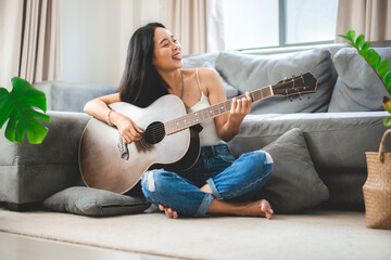 Fototapeta Asian woman playing music by guitar at home, young female guitarist musician lifestyle with acoustic art instrument sitting to play and sing a song making sound in hobby in the house room obraz