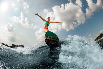 smiling young woman riding down the blue splashing wave on wakesurf at warm day