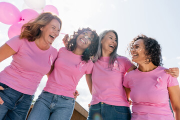 Low angle view of cheerful interracial women with pink ribbons and balloons hugging outdoors - Powered by Adobe