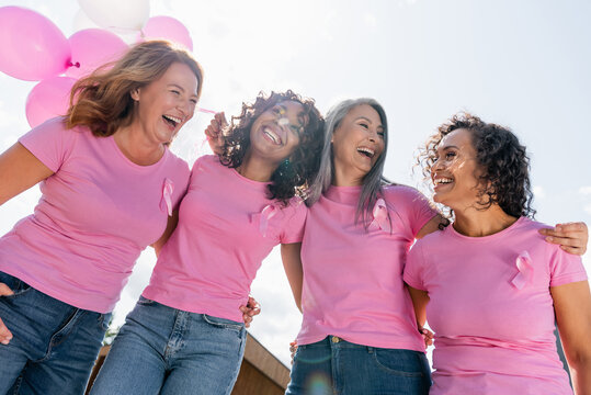 Low angle view of cheerful interracial women with pink ribbons and balloons hugging outdoors