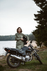 Naklejka na ściany i meble Portrait of a female biker standing outdoors. A woman holds a helmet in her hands on the background of a motorcycle.
