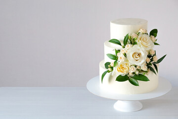 Three-tiered white wedding cake decorated with flowers and green leaves on a white wooden...