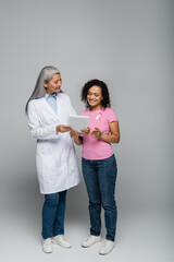 Cheerful asian doctor holding digital tablet near african american patient with pink ribbon on grey background