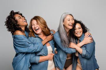 Happy multiethnic women in underwear and denim shirts embracing isolated on grey, body positive...