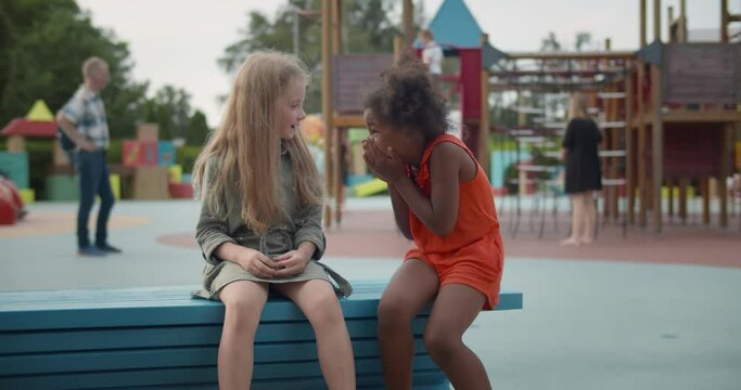 Smiling multiethnic little girls sitting on wooden bench outdoors whispering secret