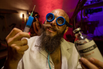 Portrait of a mad bearded doctor with a bottle of vaccine and a syringe in his hands in a dark...